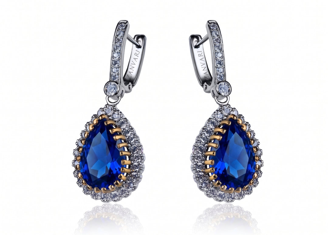 A pair of silver and gold drop earrings with teardrop-shaped blue gemstones, surrounded by small clear stones, on a white background with a soft reflection.