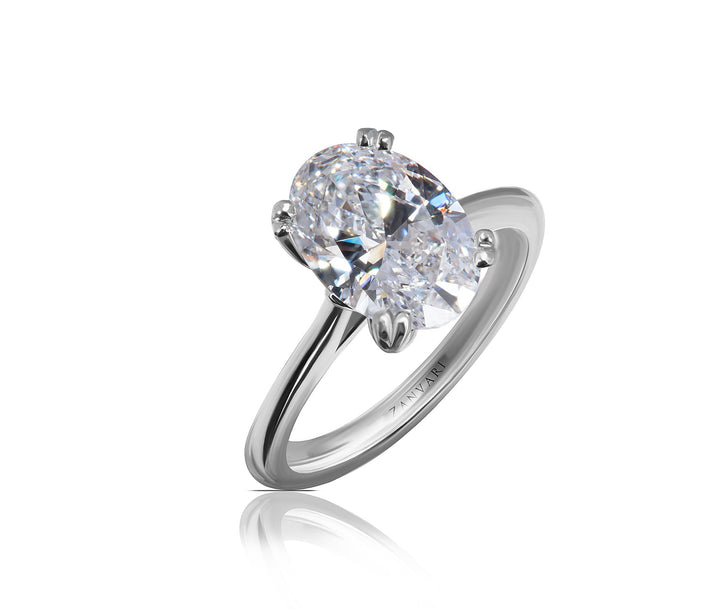 A oval brilliant-cut diamond solitaire ring in a four-prong setting on a polished silver-colored band, shown against a clean white background with a soft reflection below.