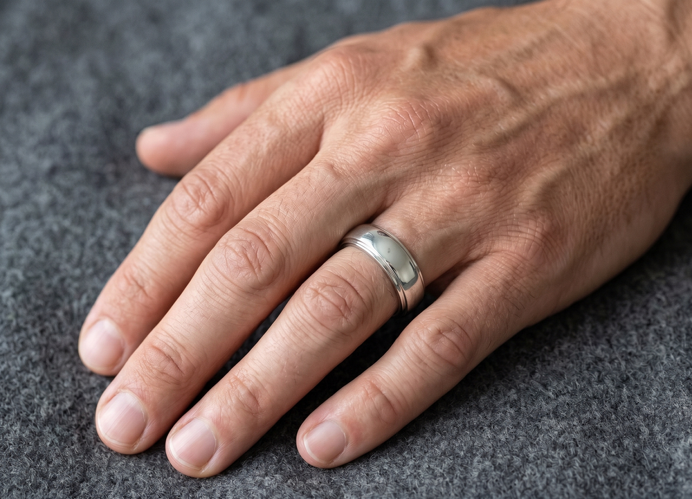 A close-up of a man's hand resting on a textured gray surface, wearing a simple polished silver band ring on one finger.