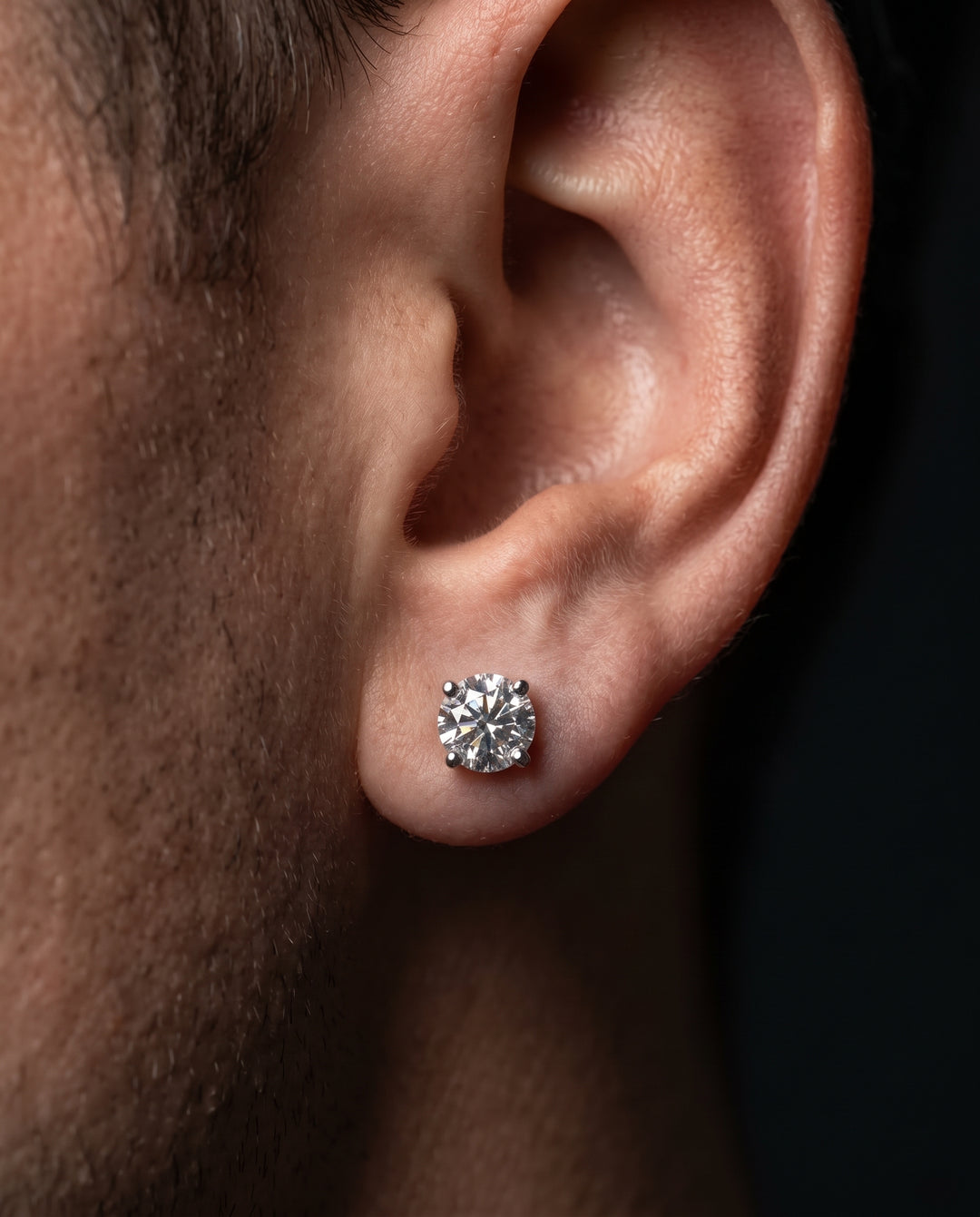 Macro side-profile photo of a man's ear wearing a sparkling round brilliant diamond stud earring set in white metal against a dark background.