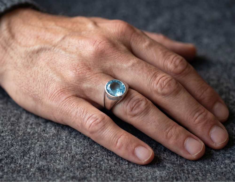 A close-up of a person's hand resting on a textured gray surface, wearing a silver ring with a round light blue topaz gemstone.