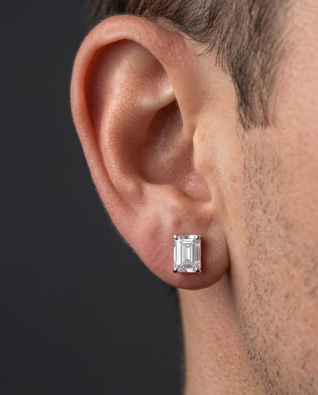 A close-up profile view of a male model's earlobe wearing a four-prong emerald-cut diamond stud earring, set against a dark neutral background under soft directional lighting.