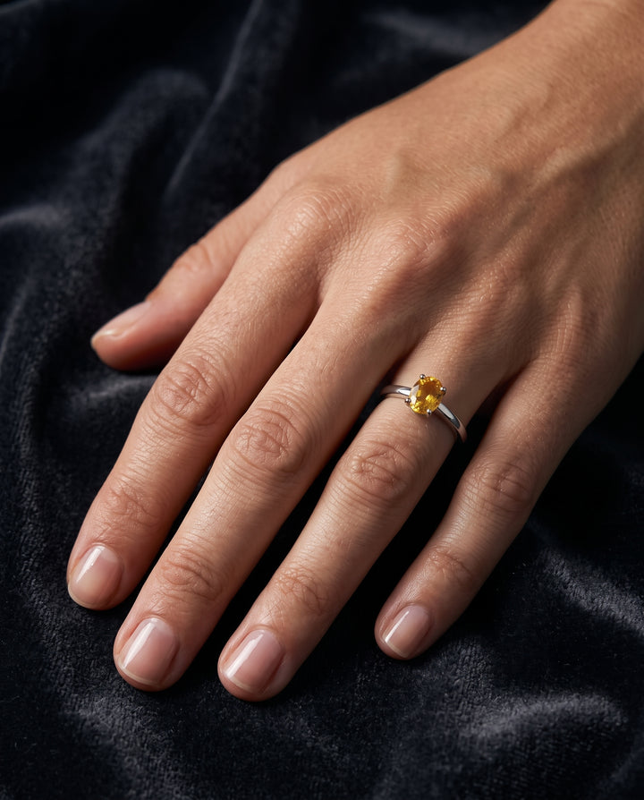 A close-up editorial photo of a woman's hand resting on dark grey velvet, wearing a slender silver solitaire ring set with a small faceted golden-yellow citrine under soft natural light.