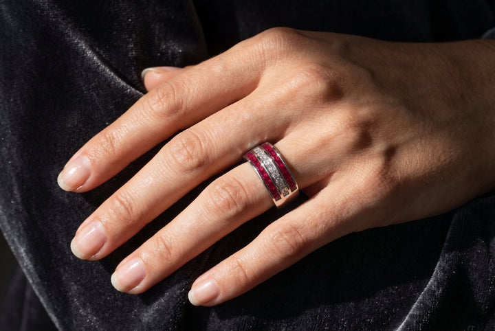 A close-up of a hand wearing a ring with a gold band set with red gemstones and clear stones, resting on a dark velvet fabric.