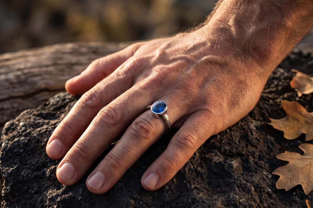 A detailed close-up photograph of a man's right hand with prominent veins and detailed skin texture, resting on a dark, porous volcanic rock in warm, natural sunlight. A polished silver bezel-set ring with a faceted oval blue sapphire is on the ring finger. To the right of the hand, three dry, brown oak leaves lie on the rock. In the background is a weathered, blurred piece of wood, all captured with a shallow depth of field.