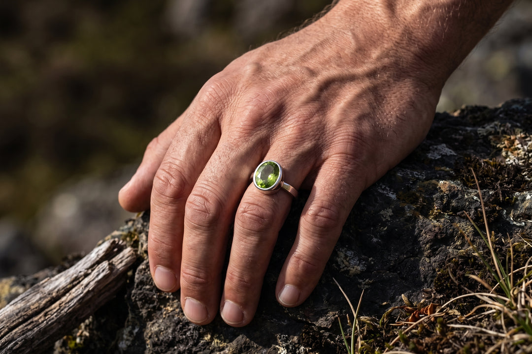 A close-up editorial shot of a strong, masculine hand resting firmly against dark, mossy rock and weathered wood outdoors. Natural sunlight highlights the hand's defined knuckles, veins, and realistic skin texture. On the middle finger is a sleek silver ring featuring a bezel-set, faceted oval peridot gemstone that glows with a vivid olive-green hue under the side lighting. The background shows a soft-focus natural landscape with earthy tones.