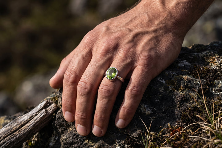 A close-up editorial shot of a strong, masculine hand resting firmly against dark, mossy rock and weathered wood outdoors. Natural sunlight highlights the hand's defined knuckles, veins, and realistic skin texture. On the middle finger is a sleek silver ring featuring a bezel-set, faceted oval peridot gemstone that glows with a vivid olive-green hue under the side lighting. The background shows a soft-focus natural landscape with earthy tones.