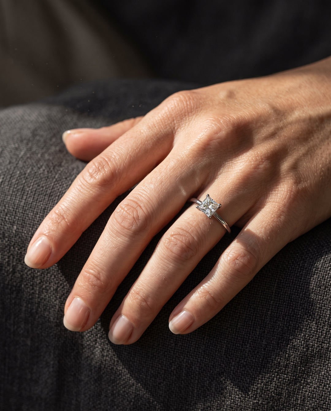 A close-up of a hand wearing a silver ring with a square-cut clear moissanite gemstone, resting on a dark textured fabric surface.