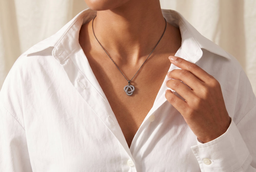 A close-up, editorial view of a person wearing an open-collared white button-down shirt and a delicate dark silver necklace featuring a small, diamond-encrusted knot pendant resting against the collarbone. One hand with manicured nails gently touches the shirt collar.
