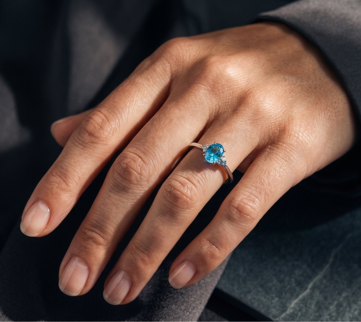 A close-up of a hand wearing a delicate silver ring with a round-cut bright blue topaz gemstone, set against a dark gray background.