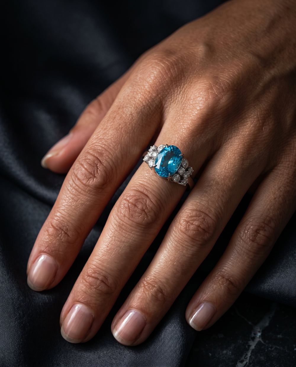 A woman's hand resting on dark fabric, wearing a platinum or white gold statement ring set with an oval vibrant blue topaz flanked by small pavé diamonds.