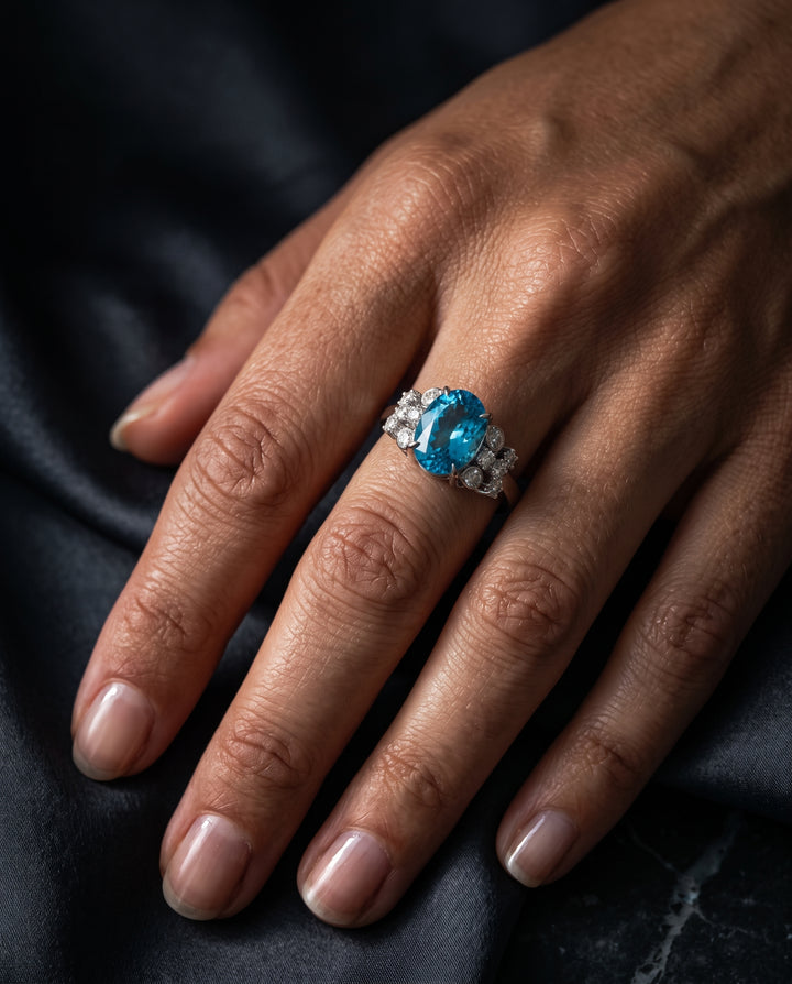 A woman's hand resting on dark fabric, wearing a platinum or white gold statement ring set with an oval vibrant blue topaz flanked by small pavé diamonds.