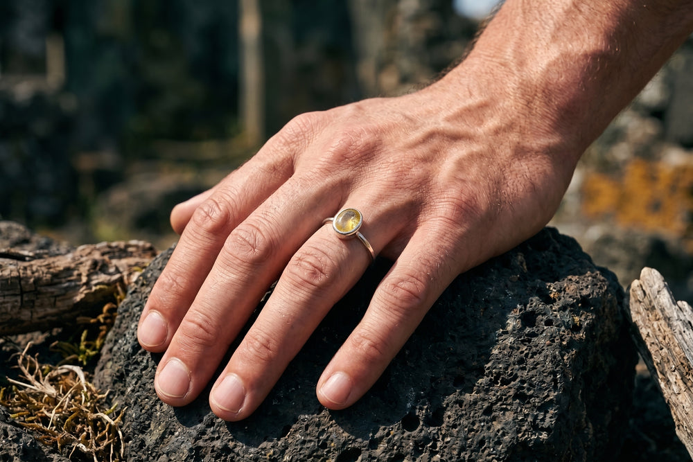 A close-up of a man's hand resting on a dark volcanic rock, wearing a silver band ring with a round yellow stone in a bezel setting.