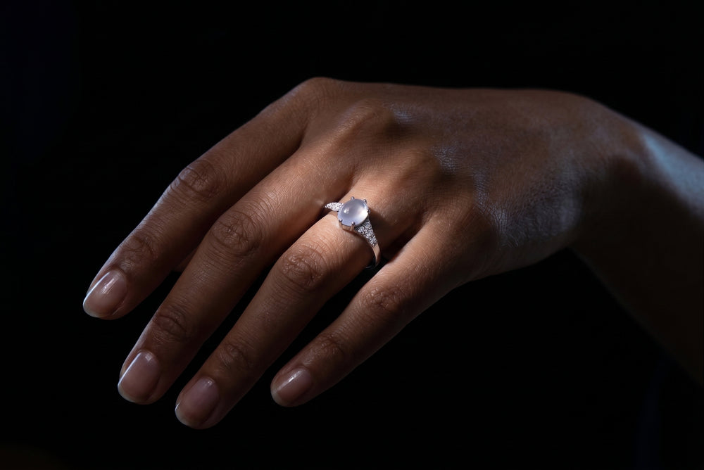 A close-up of a hand wearing a silver-toned ring with a round clear gemstone, set against a solid black background.