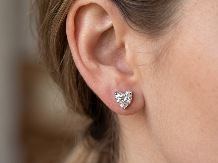 "A woman's earlobe featuring a brilliant-cut, heart-shaped moissanite stud earring set in white metal."