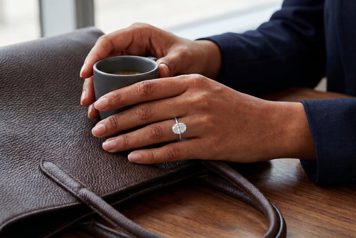 A close-up lifestyle shot of a person holding a gray ceramic mug next to a brown leather bag, wearing a silver ring with a oval diamondclear gemstone on their ring finger.