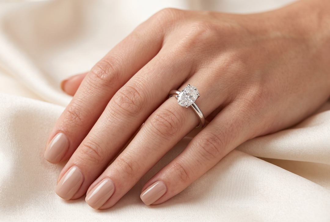 Close-up view of a woman’s hand resting on soft neutral fabric, wearing a delicate oval-cut lab-grown diamond solitaire ring in a four-prong white gold setting.