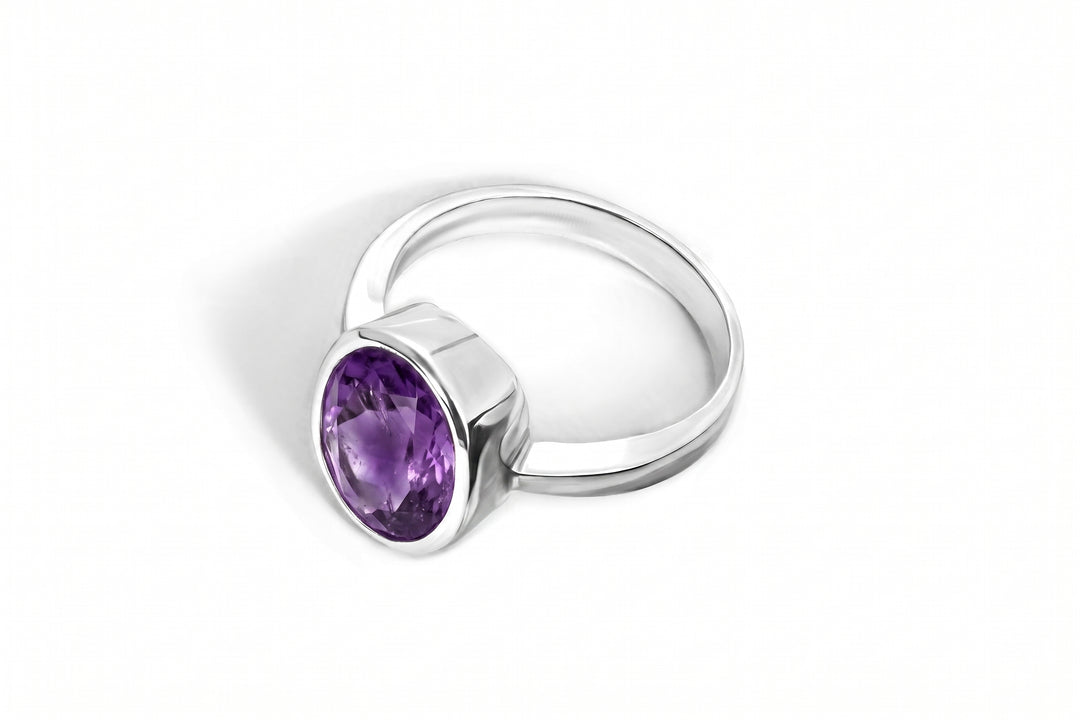 Studio product photograph of a modern silver ring centered on a clean white background. The ring features an open-band design with structured, geometric shoulders and a prominent bezel setting holding a large, faceted oval amethyst gemstone with rich purple tones.
