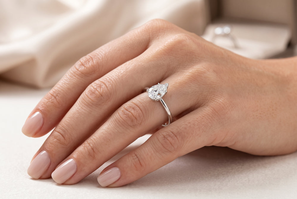 Macro close-up of a woman’s hand wearing a dainty pear-cut lab-grown diamond solitaire engagement ring on her ring finger, set in polished white gold.