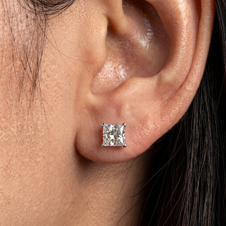 Close-up photo of a woman's earlobe wearing a small, four-prong set princess-cut diamond stud earring.