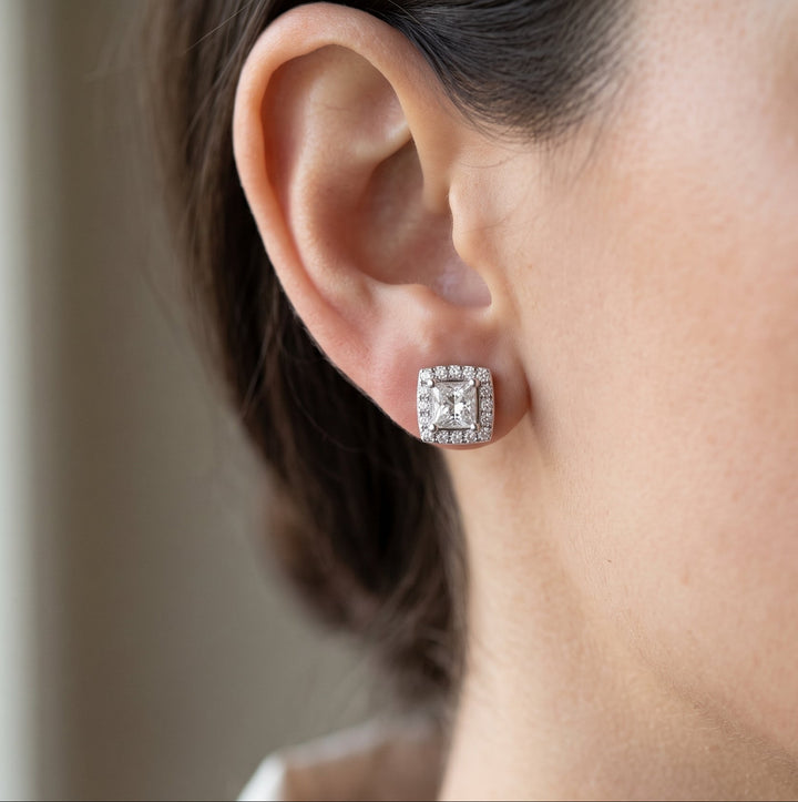 A close-up profile view of a woman with dark hair wearing a sparkling princess-cut diamond stud earring with a square halo on her earlobe.