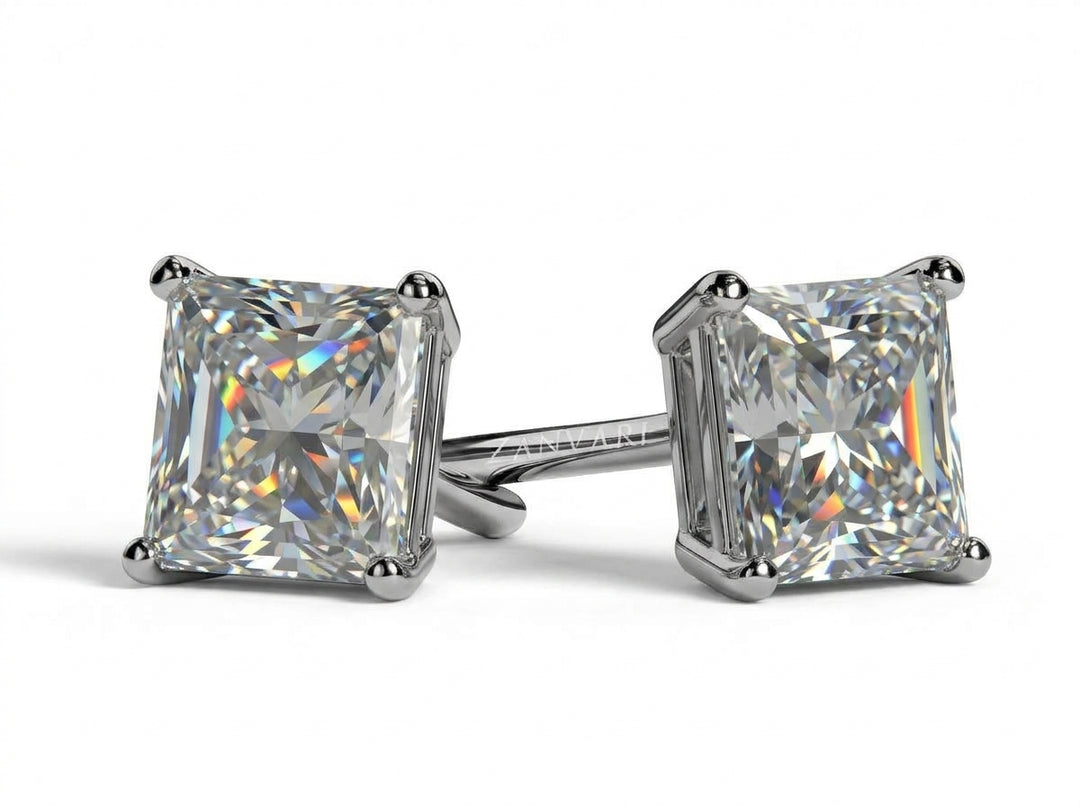 Pair of princess-cut diamond stud earrings set in four-prong mounts, displayed on a clean white surface with subtle reflections and shadows.