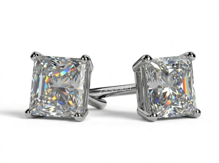 Pair of princess-cut diamond stud earrings set in four-prong mounts, displayed on a clean white surface with subtle reflections and shadows.