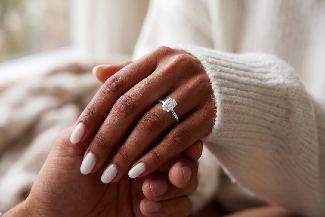 A close-up lifestyle shot of a person's hand resting on another person's hand, with a silver engagement ring featuring a oval diamond clear gemstone worn on the first person's ring finger. The person is wearing a cream-colored ribbed knit sweater.