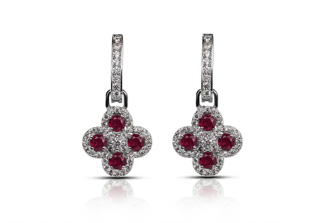 A pair of silver-tone dangle earrings featuring a four-petal flower design set with round red gemstones and surrounded by small clear pavé stones, shown on a white background with a soft reflection.
