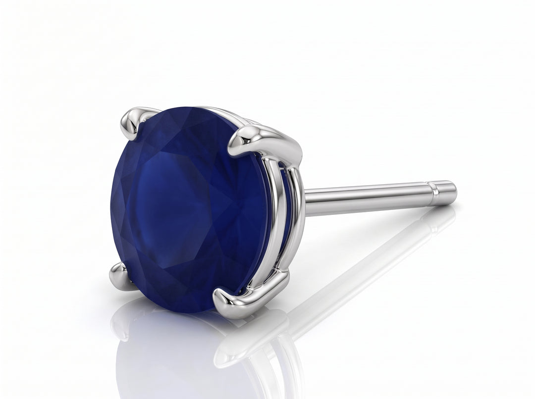 A single round-cut deep blue sapphiregemstone stud earring in a polished silver-toned prong setting, shown on a clean white background with a soft reflection.