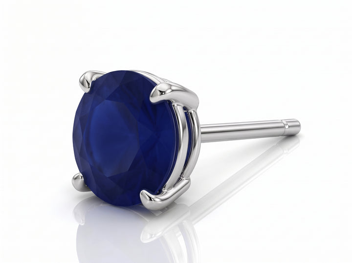 A single round-cut deep blue sapphiregemstone stud earring in a polished silver-toned prong setting, shown on a clean white background with a soft reflection.