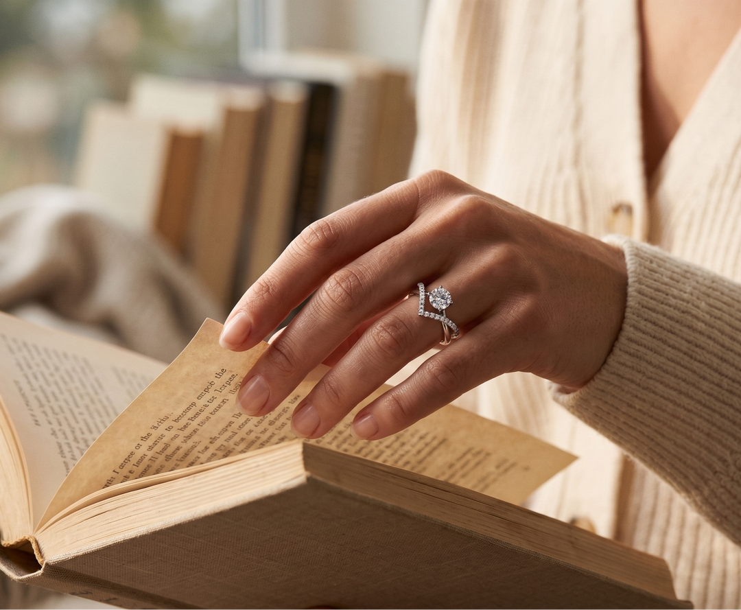 A close-up lifestyle shot of a person's hand wearing a silver ring with a large round clear moissanite gemstone and smaller side stones, while gently turning a page of an open book. The person is wearing a cream-colored ribbed cardigan sweater.
