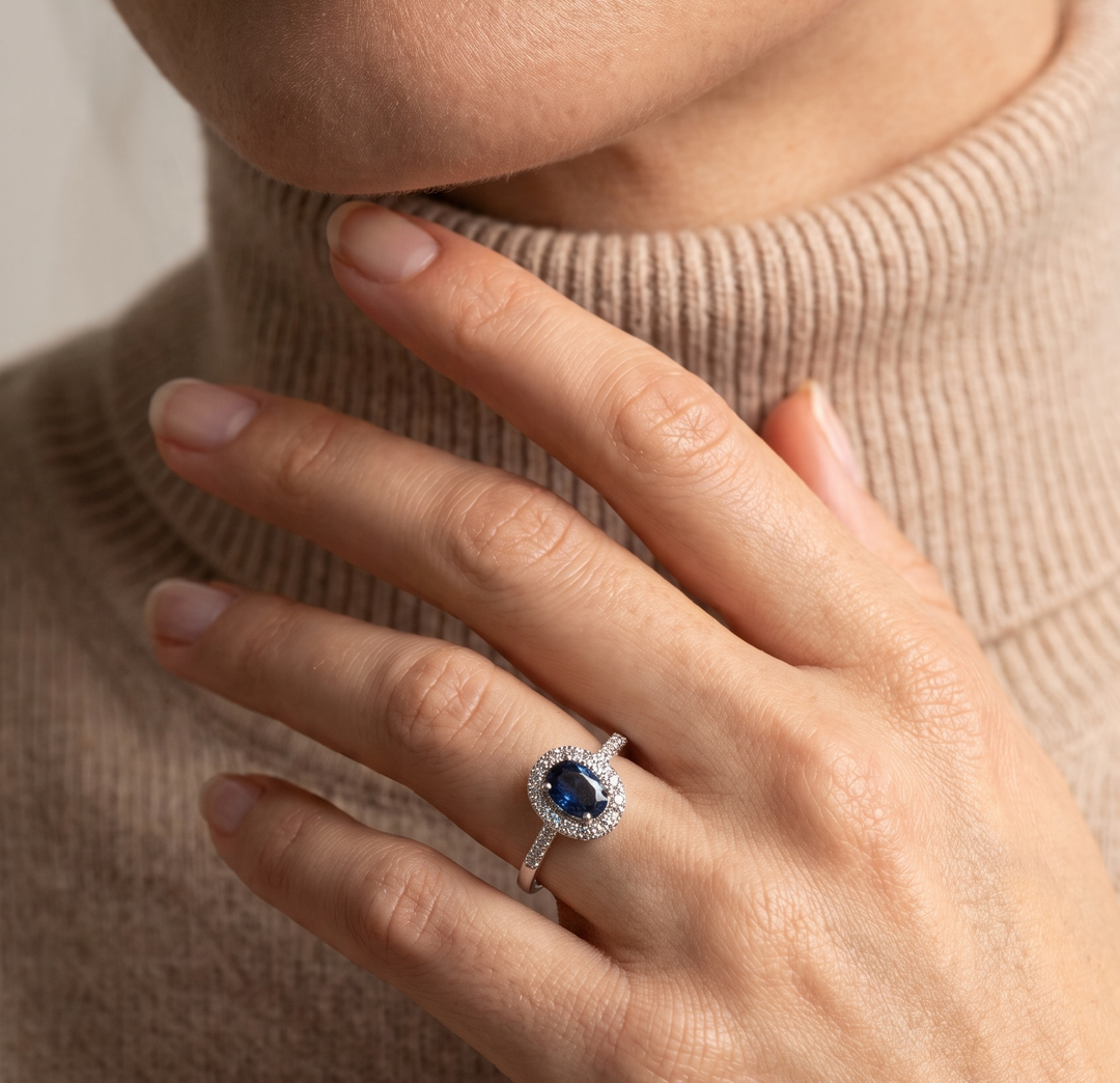 A close-up lifestyle shot of a model wearing a silver ring with a round-cut blue sapphire and a halo of small clear stones, set against a beige ribbed turtleneck sweater.