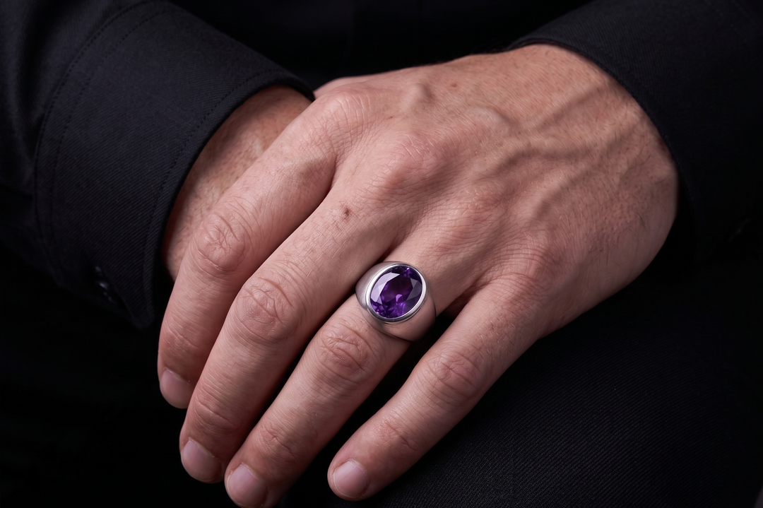 A silver ring with a large oval purple amethyst gemstone worn on a man's finger, shown against a dark black shirt background.