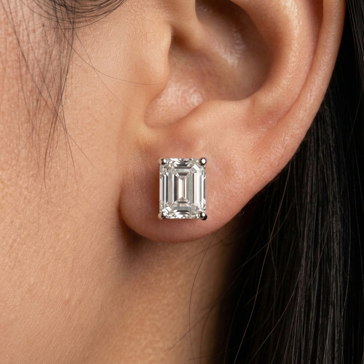Close-up of a woman's earlobe wearing a four-prong, emerald-cut diamond or moissanite stud earring in a white metal setting.