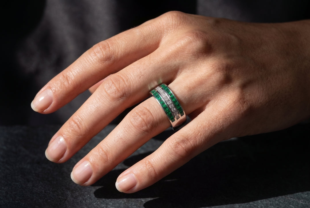 A close-up of a hand wearing a silver ring with a double row of green and clear stones, set against a dark, soft-focus background.