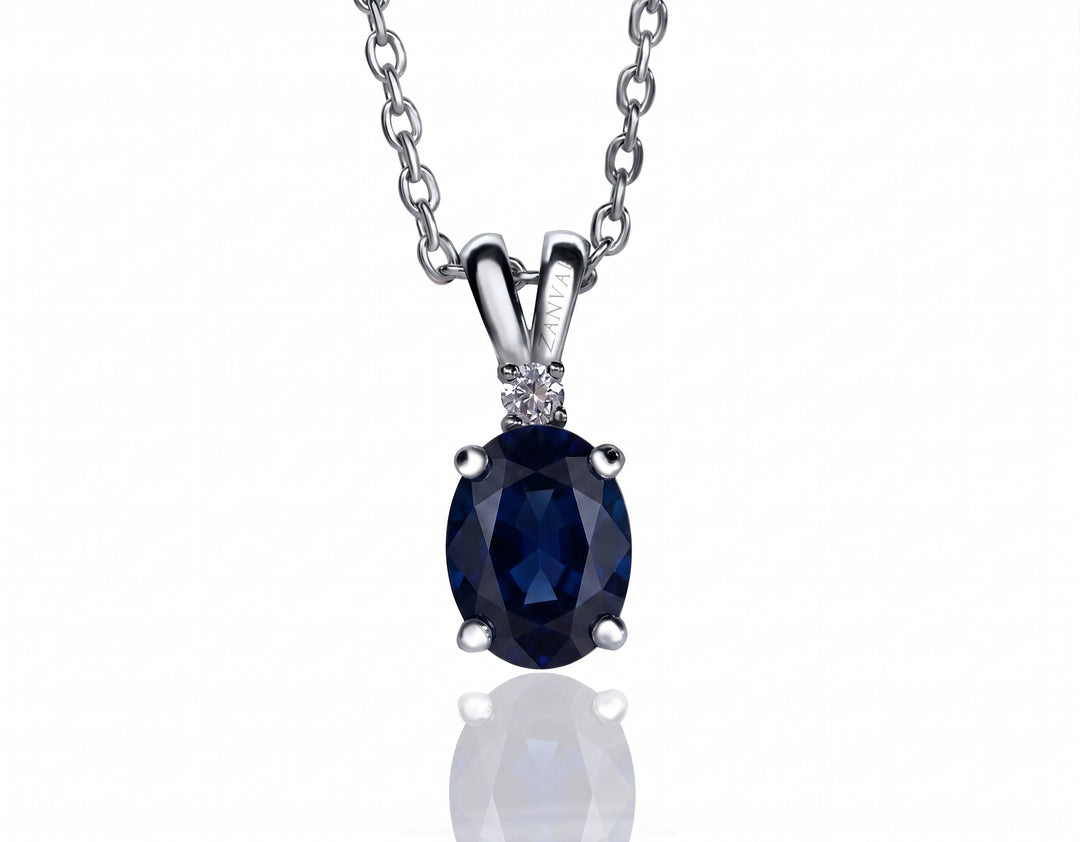 An oval-cut dark blue sapphire pendant necklace in a silver four-prong setting, suspended from a silver link chain against a white background with a soft reflection below.
