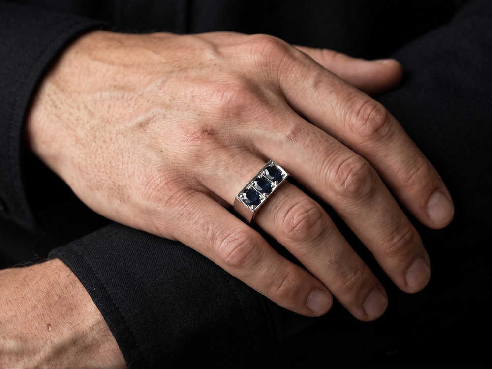 A close-up of a man's hand wearing a silver ring with three dark blue oval sapphire gemstones, set against a dark black clothing background.