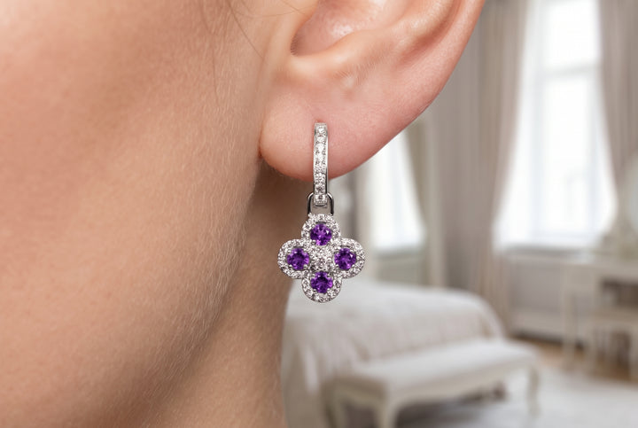 A close-up of a person's ear wearing a silver-toned huggie hoop earring with a four-petal flower charm set with purple amethysts and small clear stones.