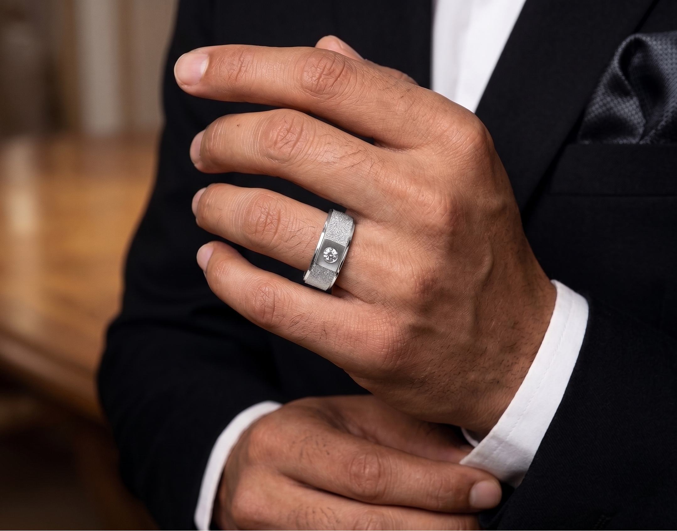 Hand wearing a silver ring with a Moissanite, against a blurred background of a person in formal attire.