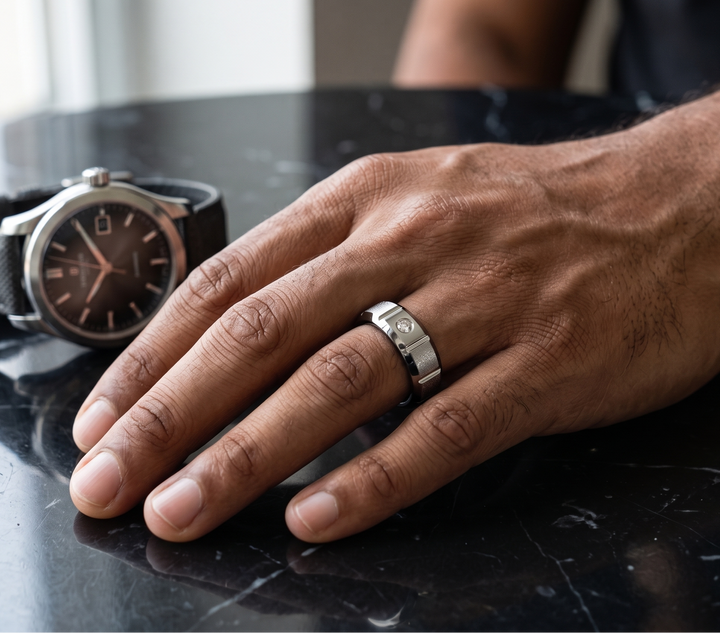A silver-toned metal band ring worn on a man's ring finger, shown next to a black leather strap wristwatch on a dark reflective tabletop.