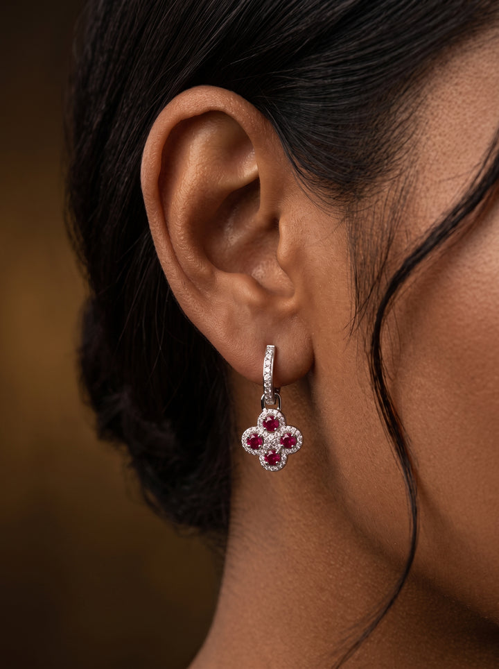 A close-up of a model's ear wearing a small dangling flower-shaped earring with four pink gemstones and a halo of clear stones in a silver-toned setting, against a warm brown background.